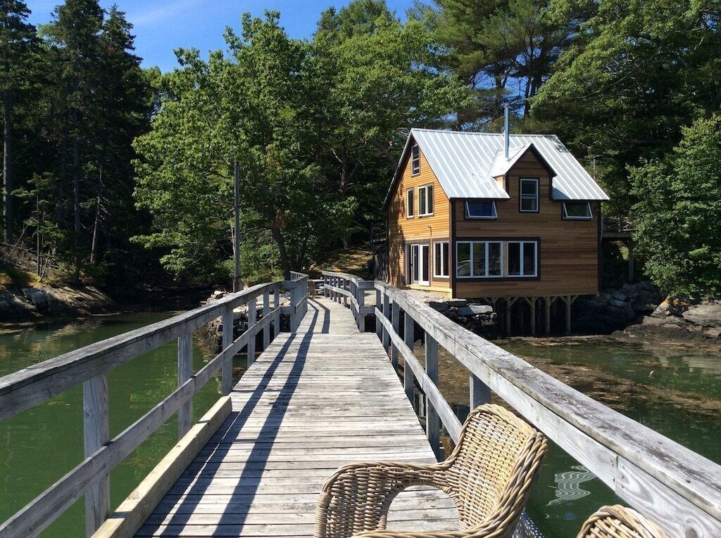 A wooden dock leading to a house with a metal roof, surrounded by trees and a body of water.