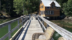 A wooden dock leading to a house with a metal roof, surrounded by trees and a body of water. - Historic Boathouse over Water, Stunning View, Private Pier + Dock + Mooring (Westport)