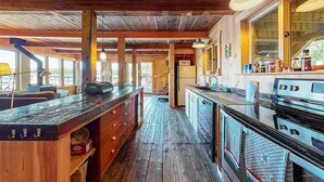 A rustic cabin interior with a long wooden counter, a stove, and a window overlooking a natural landscape. - Historic Boathouse over Water, Stunning View, Private Pier + Dock + Mooring (Westport)