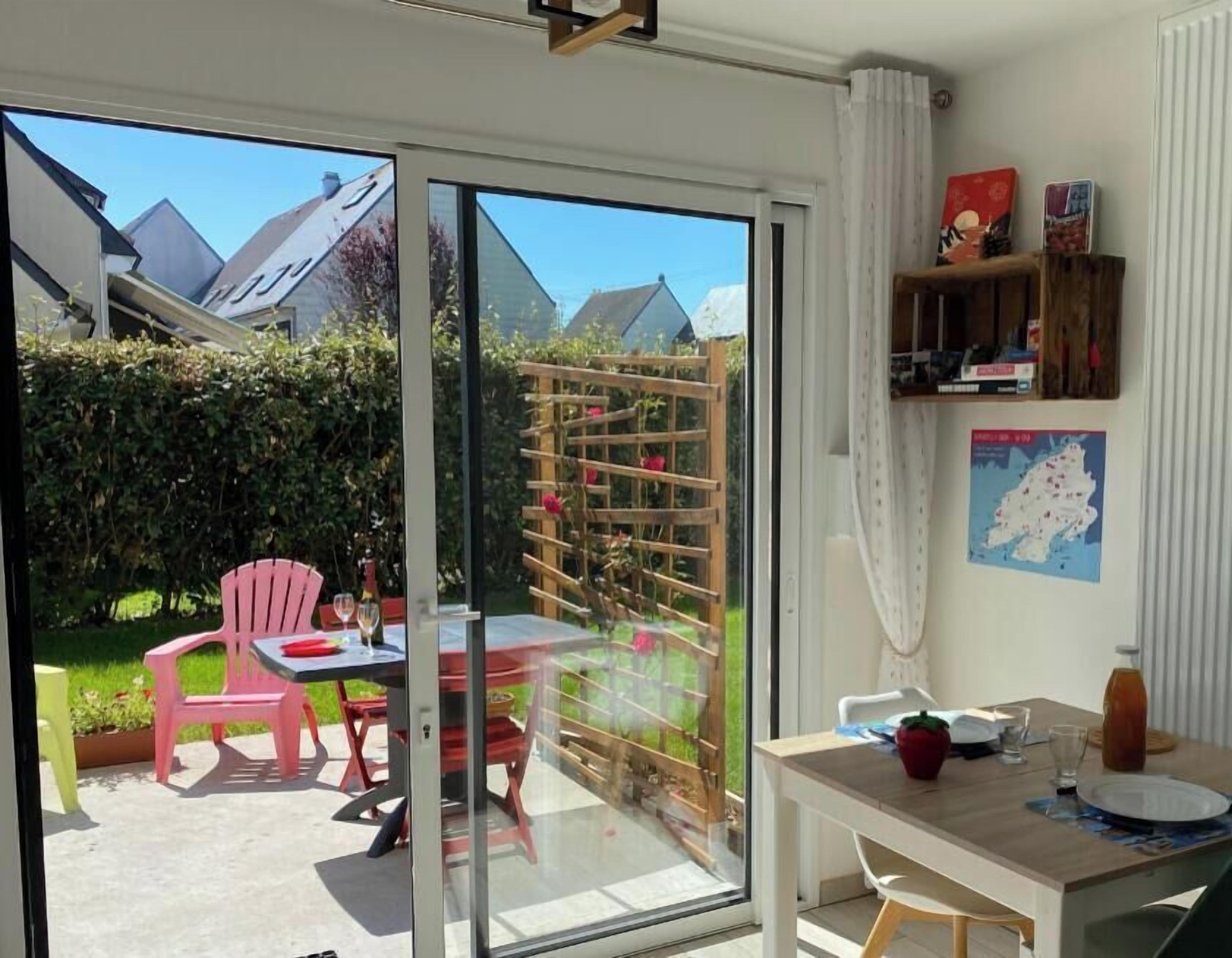 Una habitación con puerta corrediza de vidrio que da a un jardín, una mesa con sillas y un pequeño estante con objetos.