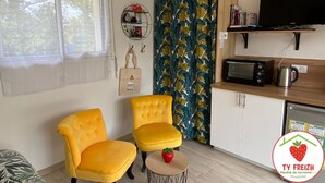 Een knus leefruimte met twee gele fauteuils, een kleine ronde tafel met een rood voorwerp, een magnetron en een televisie op een houten plank.