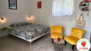 Een slaapkamer met een bed, twee gele fauteuils, een klein rond tafeltje met een rood voorwerp, een raam met zonwering en wanddecoraties, waaronder een ingelijst beeld en een plank met spullen.