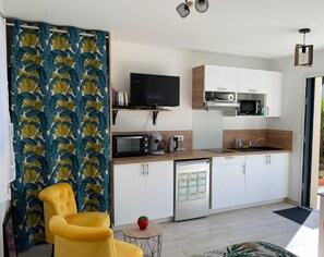 Una cocina moderna con armarios blancos, microondas y cafetera. Se ve un televisor sobre una repisa de madera y una silla amarilla.