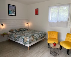Un dormitorio con una cama, dos sillones amarillos, una pequeña mesa redonda con un objeto rojo, una ventana con persianas y adornos en la pared, incluyendo un cuadro enmarcado y un estante con objetos.