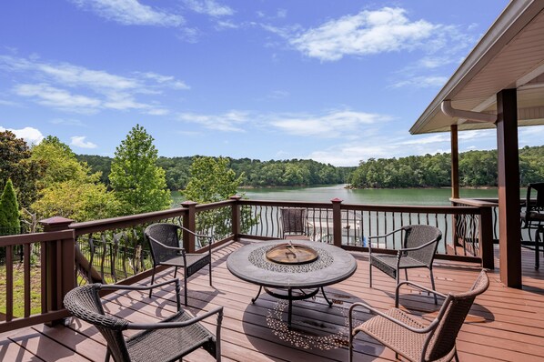 A deck with a round table and chairs, overlooking a lake and forested hills.