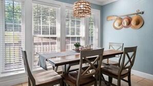 Un coin repas avec une table et des chaises, de grandes fenêtres équipées de stores, un luminaire suspendu et un porte-chapeaux fixé au mur.