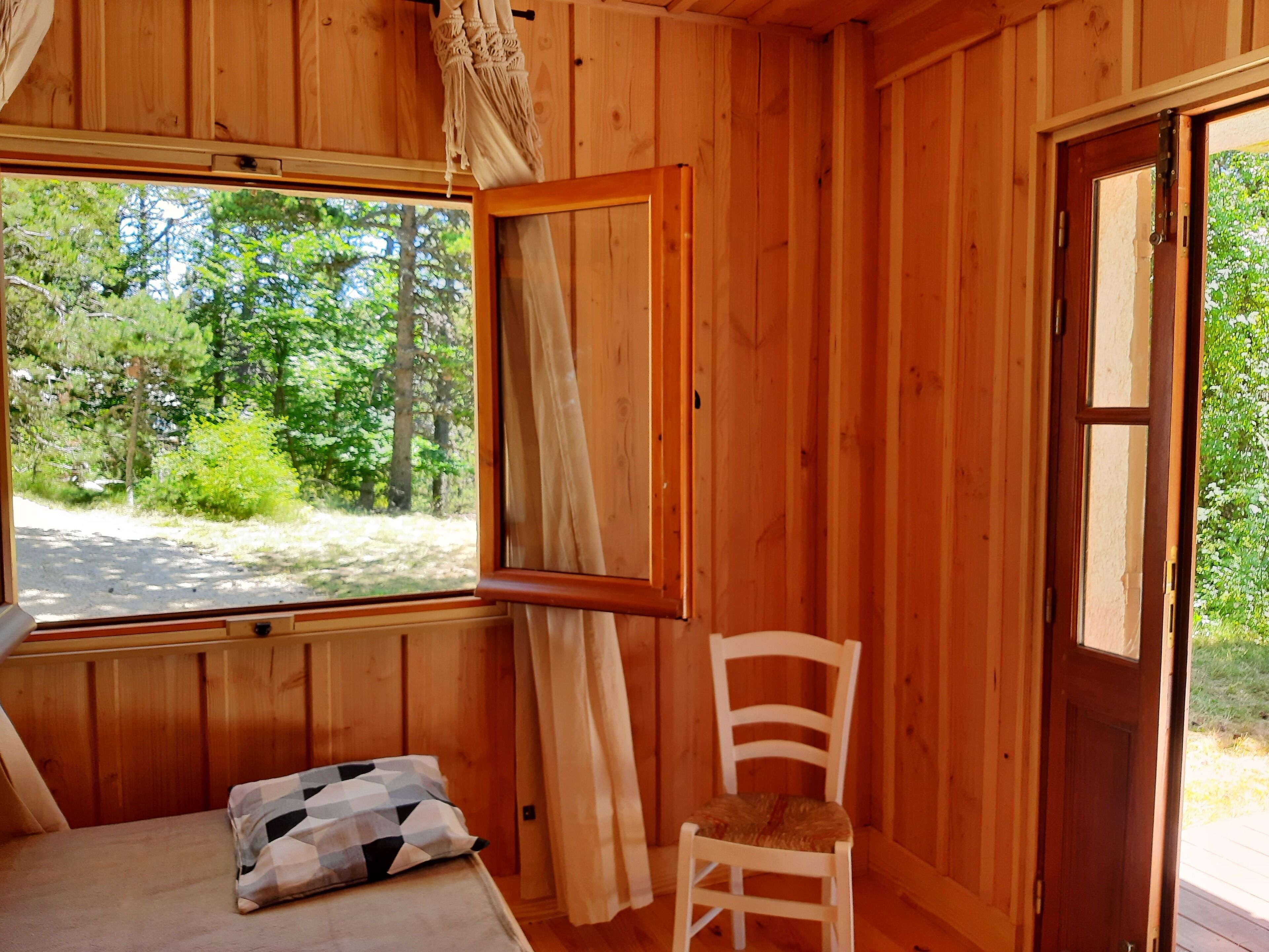 A wooden cabin room with a bed, a chair, and a window offering a view of the forest.