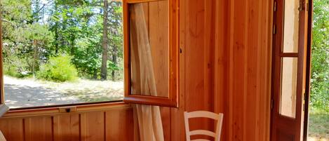 A wooden cabin room with a bed, a chair, and a window offering a view of the forest.