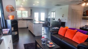 Et moderne køkken med hvide skabe, en sort bordplade og en indbygget ø. Der er et spiseområde med en sort lædersofa og orange hynder.