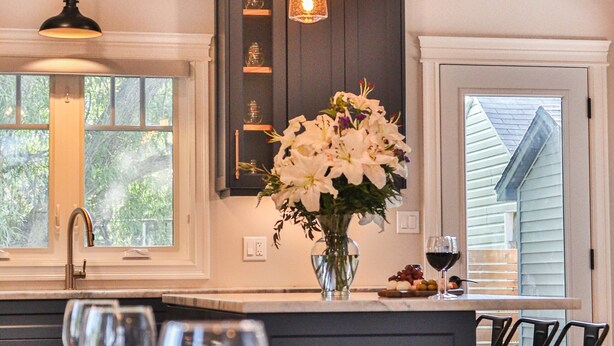 A modern kitchen with a dining area. It features a large island with a vase of flowers, a built-in wine rack, and pendant lighting. - Luxury stay at a Beach House • Sleeps 8 • Walk to Bay Beach (Crystal Beach)
