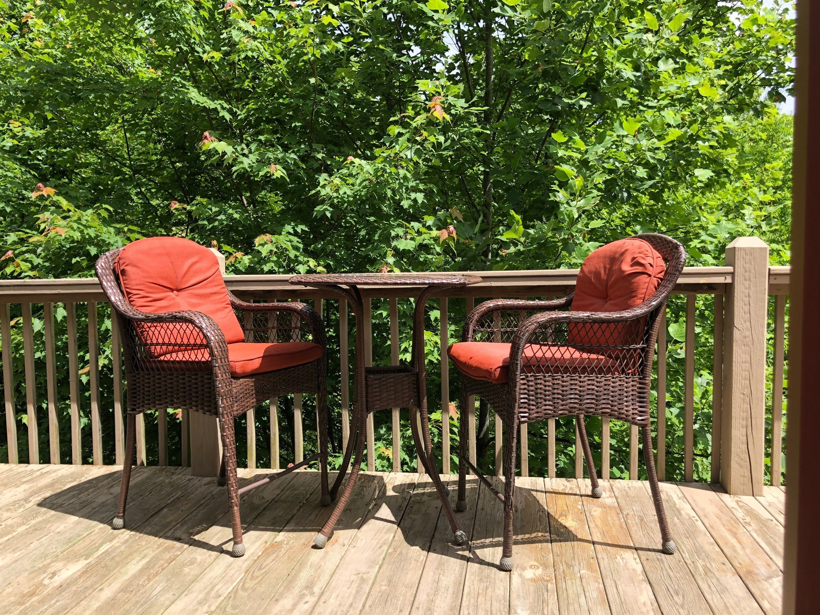 A wooden deck with wicker chairs and a table, surrounded by a wooden railing and lush green foliage.