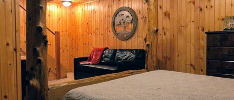 Una habitación acogedora con paredes de madera, una cama con colcha, un sofá de cuero con una manta roja y un aparador.
