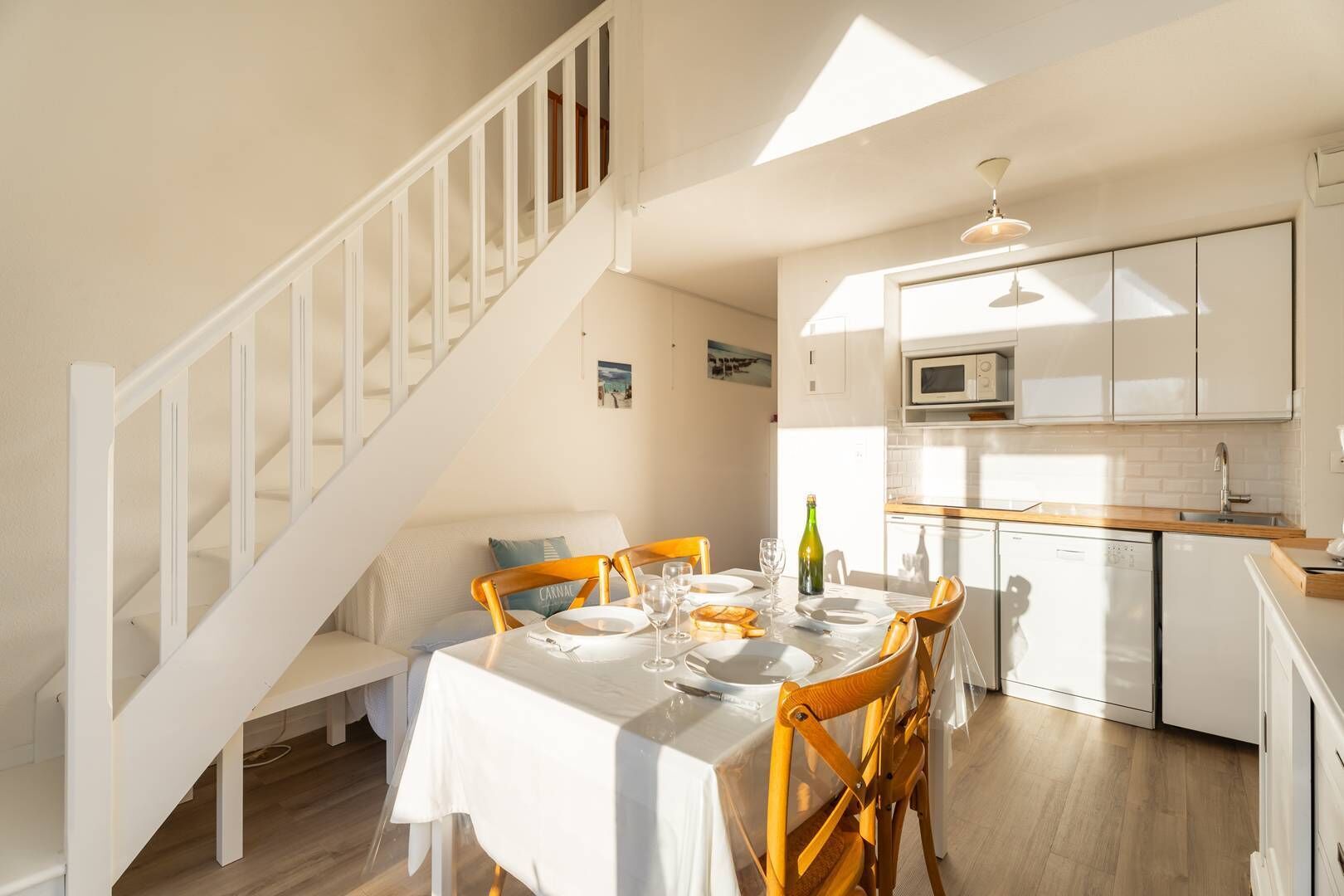 A dining area with a table set for four, a staircase, and a kitchen with white cabinets and a microwave.