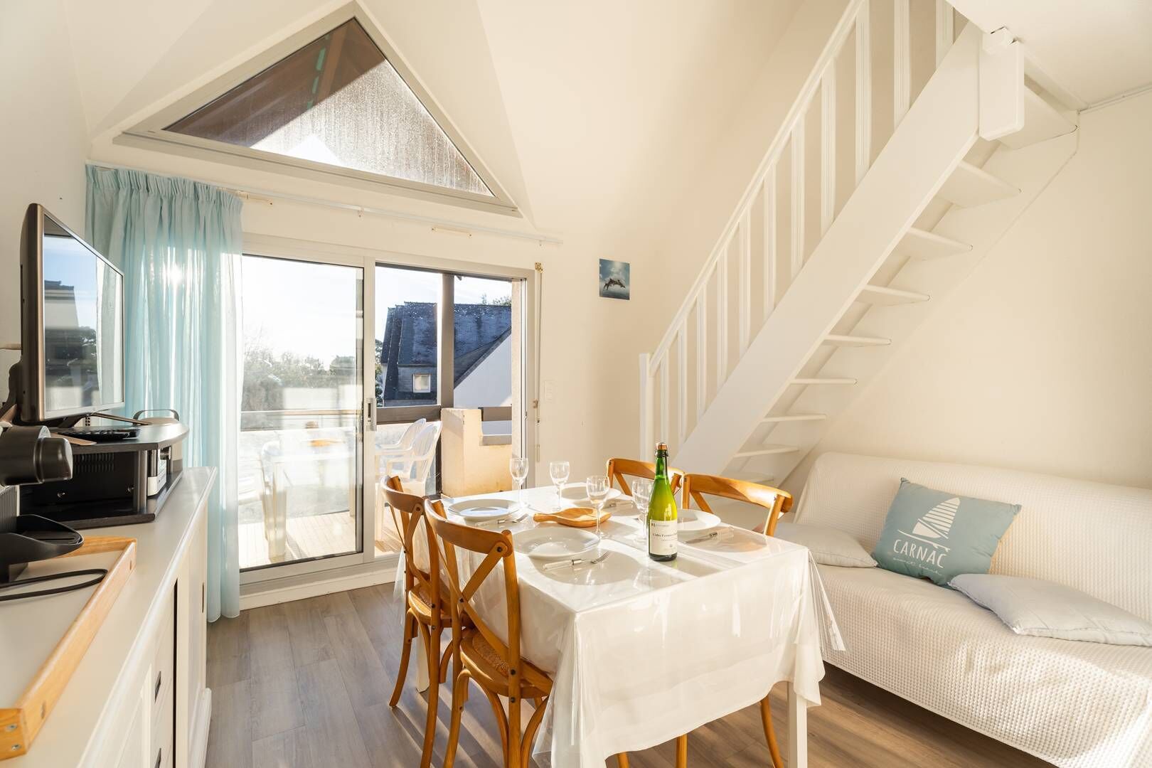 A dining area with a table set for two, a bottle of wine, and a sofa with a cushion.