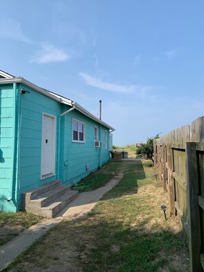 Une maison turquoise de plain-pied, avec une porte et des fenêtres blanches, un chemin en béton menant à l’entrée et une clôture en bois à l’arrière-plan.