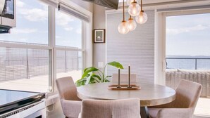 Un espace repas moderne avec une table ronde, quatre chaises, une plante en pot et une vue sur la mer grâce à de grandes fenêtres.