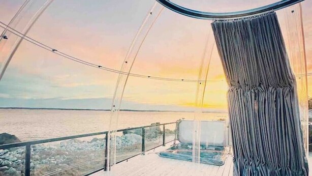 Il s’agit d’une pièce moderne, de forme circulaire, offrant une vue panoramique sur la mer au coucher du soleil. Elle comprend une terrasse en bois aux lignes courbes, un coin salon avec un canapé et une table basse, ainsi qu’une grande fenêtre agrémentée de rideaux.