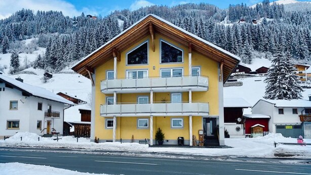 Un immeuble jaune à plusieurs étages, doté de balcons, entouré d’arbres couverts de neige et avec une montagne en arrière-plan.