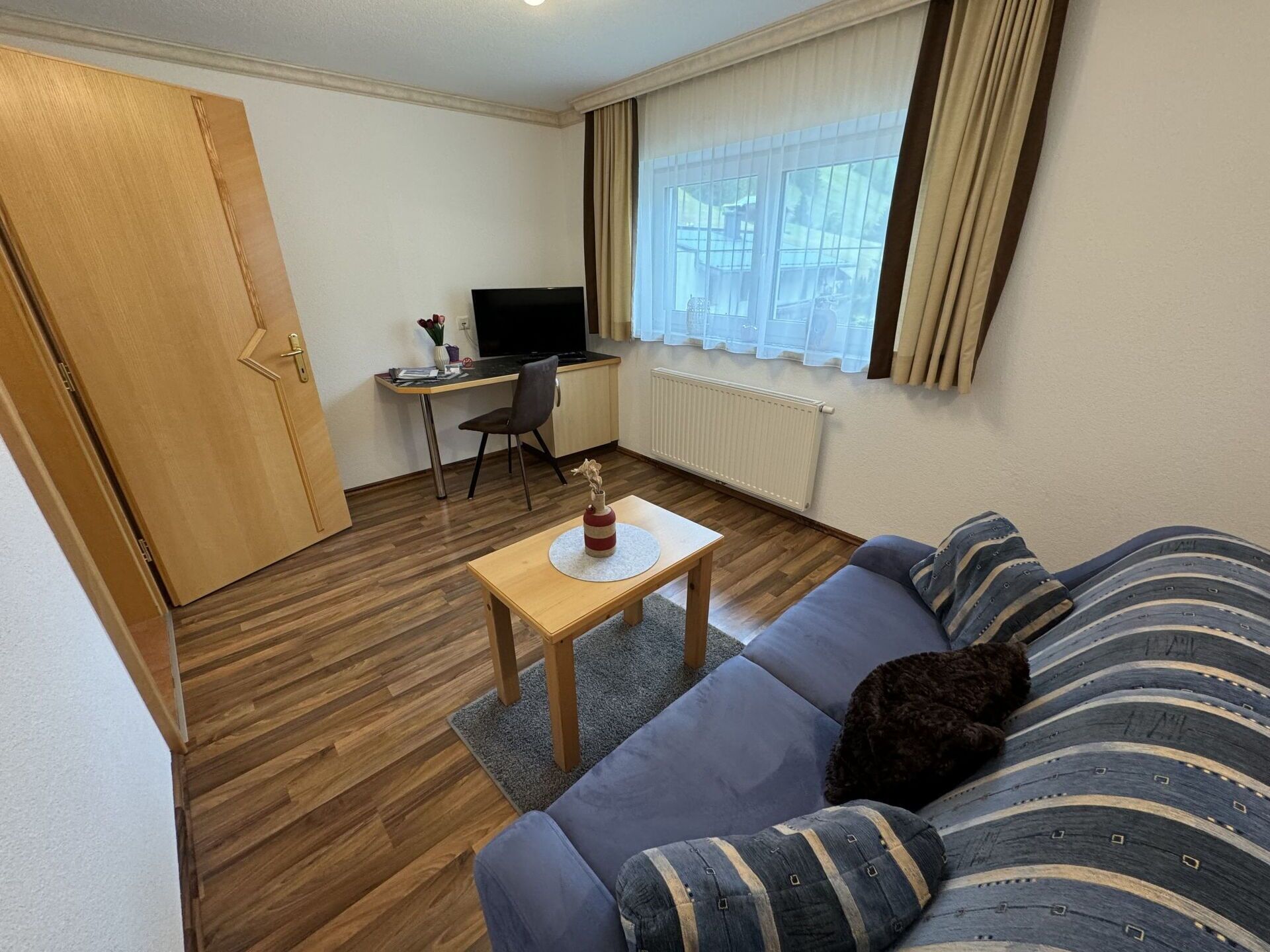 A compact living space with a sofa, a small table, a desk with a chair, a television, and a window with curtains.