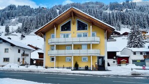 Un edificio amarillo de varios pisos con balcones, rodeado de árboles cubiertos de nieve y una montaña al fondo.