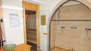 Un baño con un mueble de madera, un lavabo de vidrio verde, una ducha con pared de azulejos de piedra y la puerta de un sauna.