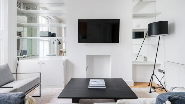 Ein modernes Wohnzimmer mit einem an der Wand montierten Flachbildfernseher, einem schwarzen Couchtisch mit einem Buch, einem grauen Sofa und einer Stehlampe.