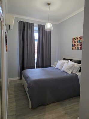 Une chambre avec un grand lit, des rideaux gris, un luminaire suspendu et une œuvre d’art colorée accrochée au mur.