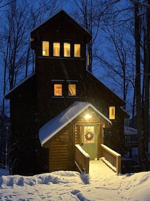 Una baita di legno con veranda e ingresso illuminato, circondata da neve e alberi.