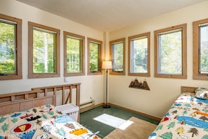 Ein Zimmer mit zwei Betten, einem Schreibtisch, einer Lampe und einem Fenster mit Blick auf Bäume.