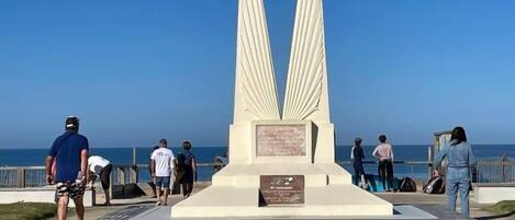 A monument with two large wings, surrounded by a paved walkway, with a clear view of the ocean in the background.