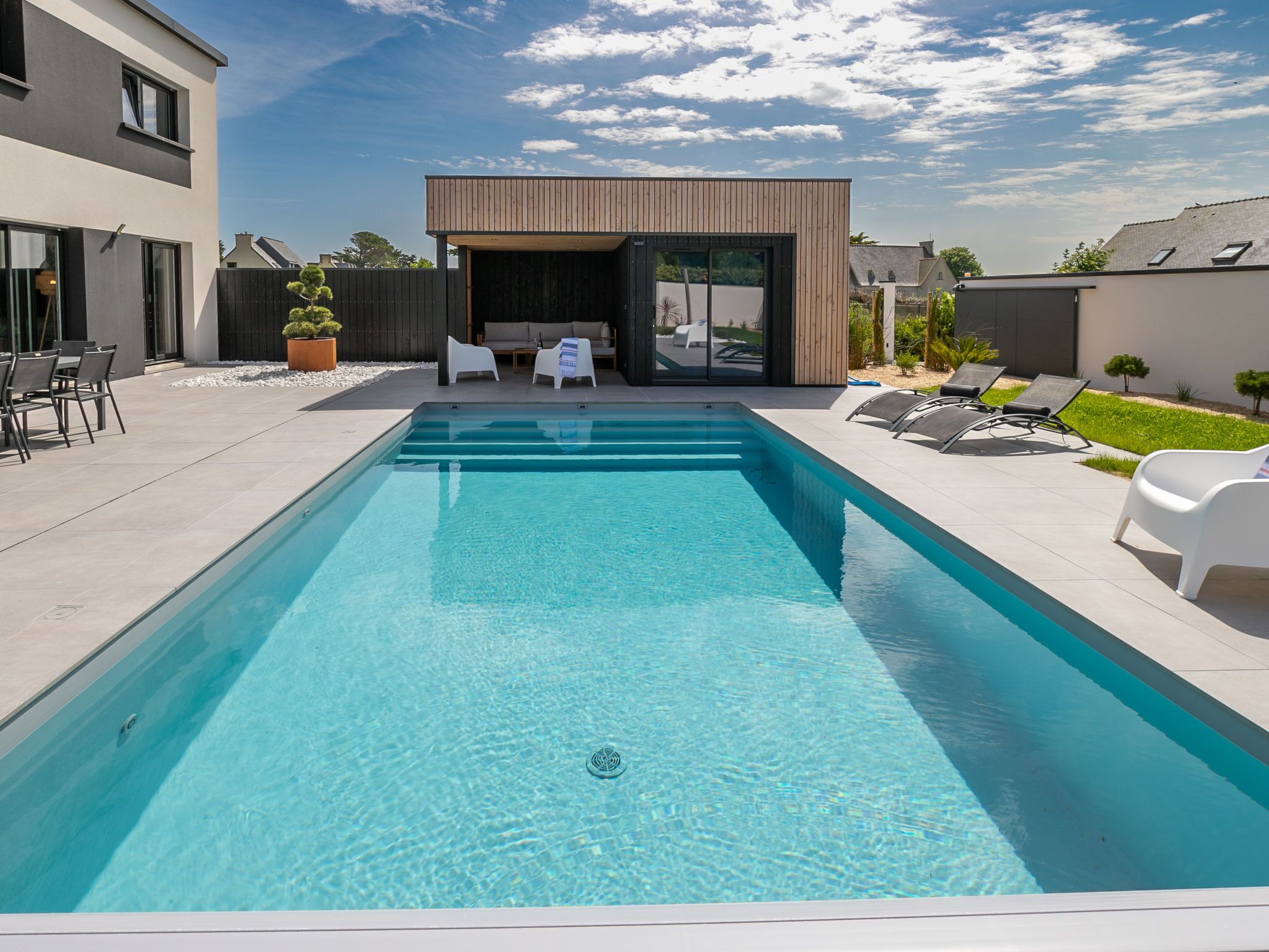 Une piscine rectangulaire à l’eau d’un bleu cristallin, entourée d’une terrasse agrémentée de chaises longues blanches et d’un espace salon extérieur moderne.