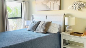 Een slaapkamer met een bed, nachtkastje, lamp, plafondventilator, raam met gordijnen en een aan de muur hangend schilderij.