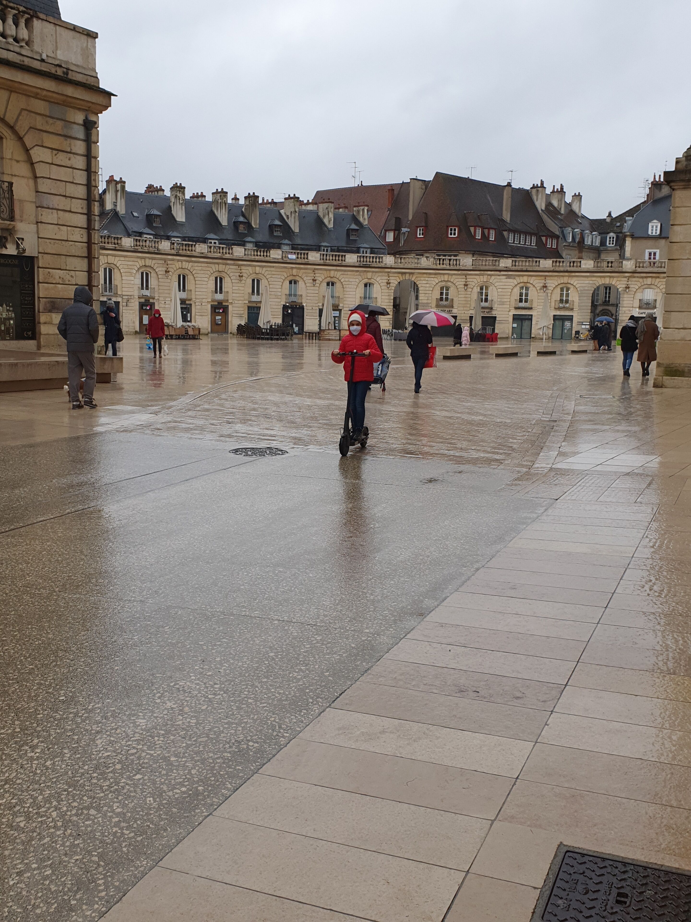 Une place spacieuse, avec un sol mouillé, des bâtiments historiques et des passants.
