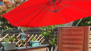 An outdoor patio with a wooden table and chairs, a red umbrella, and potted plants.