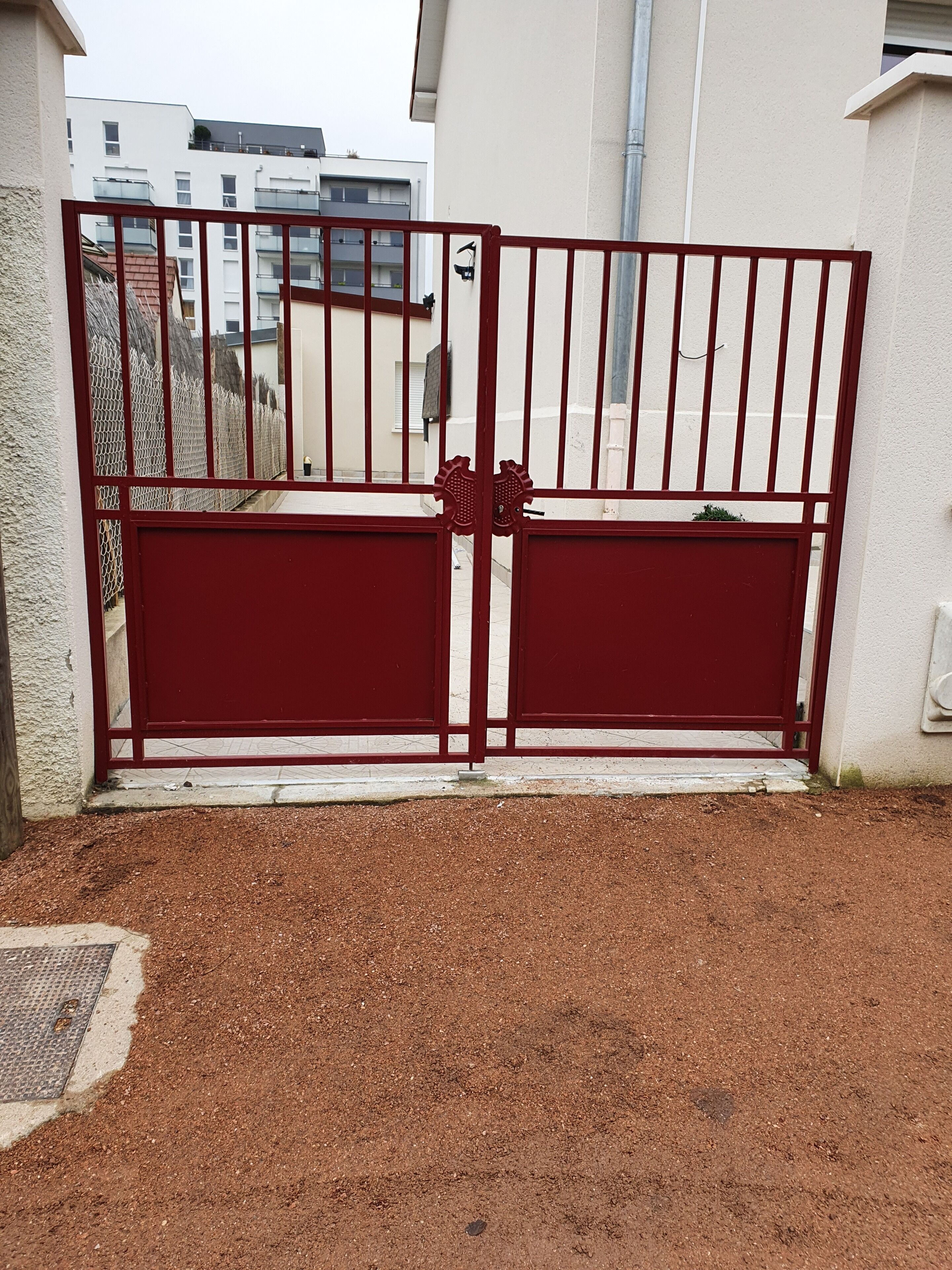 Une porte en métal rouge ornée d’un trou de serrure décoratif, qui donne sur une zone pavée surmontée d’une grille en métal.