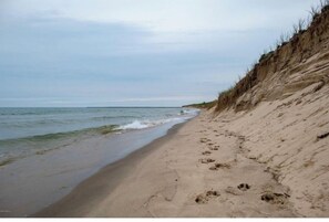 A sandy beach with footprints leading to the water, a grassy cliff, and a cloudy sky. - Lake Michigan home with beautiful  beach frontage accessible from back yard.  (West Olive)