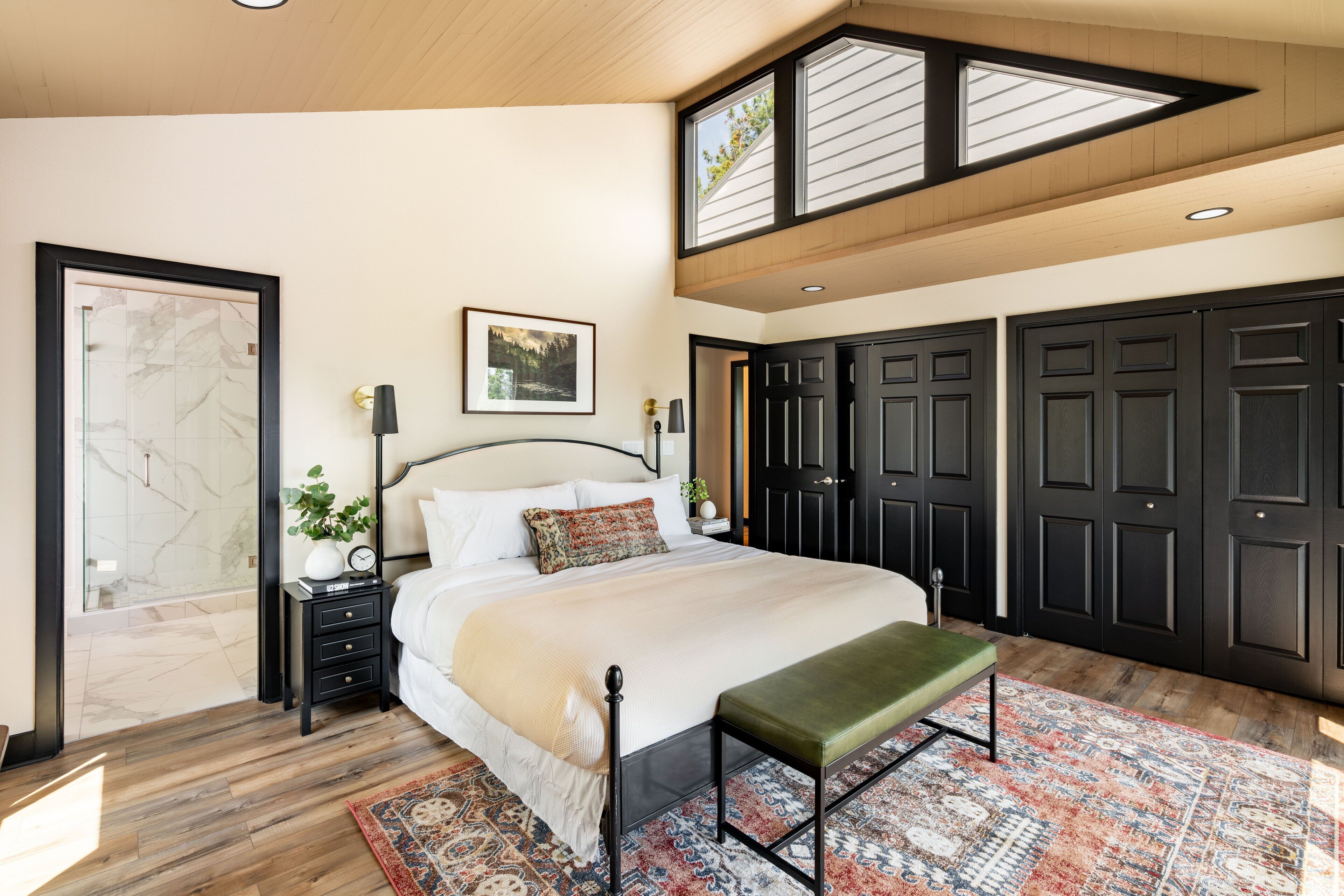 Une chambre à coucher avec un grand lit, un banc, une table de chevet, une lampe fixée au mur, un tableau encadré et une porte avec un panneau de verre.