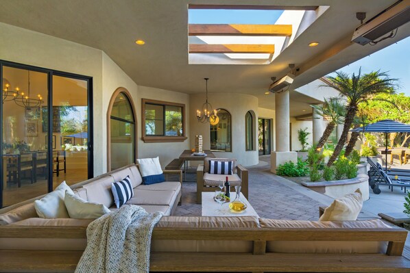 A sunlit outdoor seating area with a sofa, chairs, and a table set for dining, overlooking a landscaped garden with palm trees.