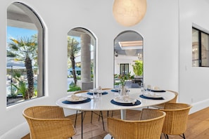 Comedor con mesa redonda, seis sillas de mimbre y vista a una piscina y palmeras a través de amplios ventanales.