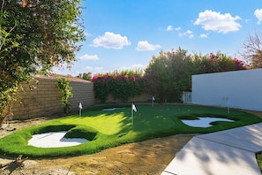 Un campo de mini golf con un césped verde para los putts, zonas de arena y una pared blanca al fondo.