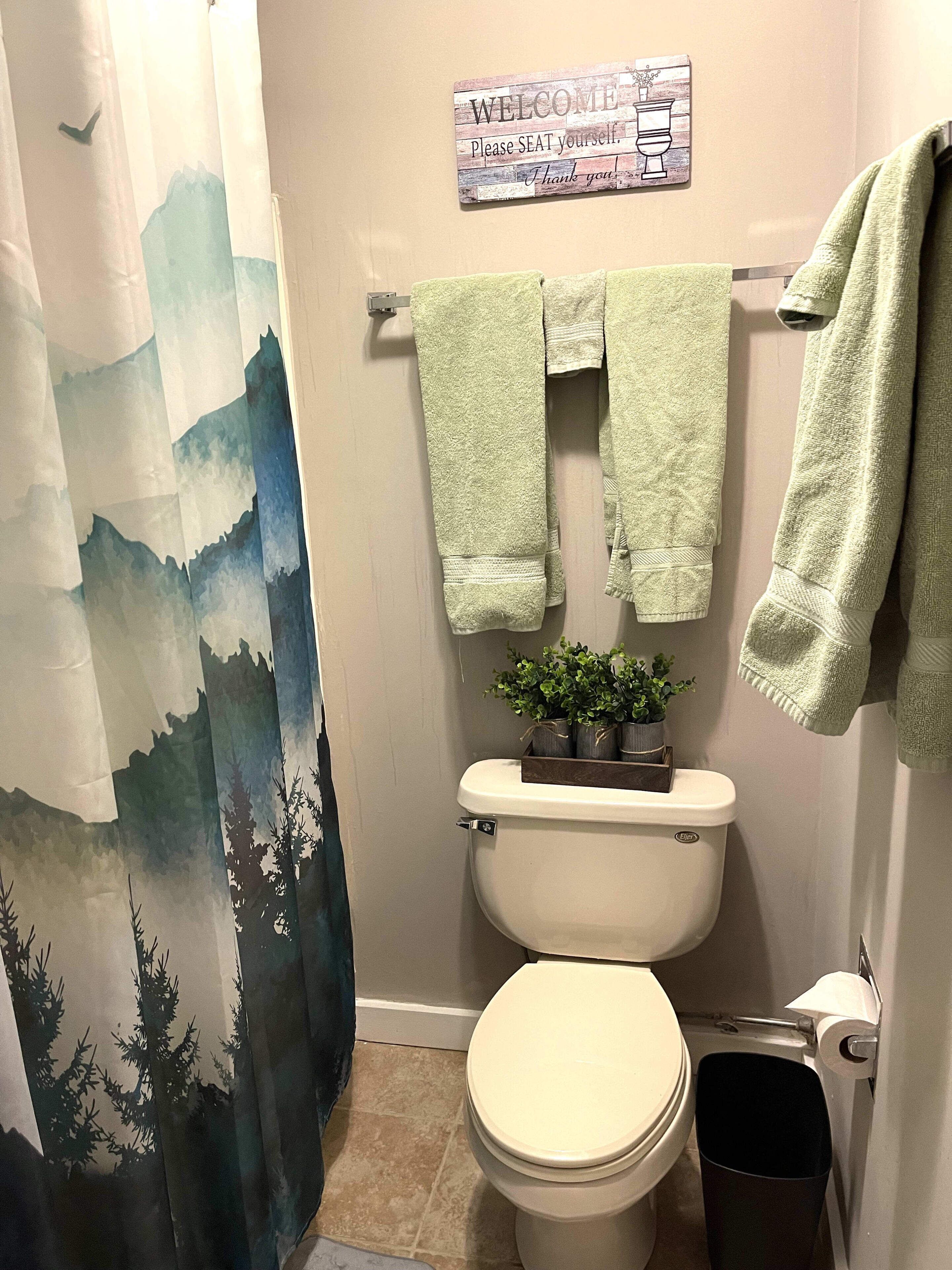 Un bagno con il water, un portaservizi verde, un cartello di benvenuto e una tenda da doccia a tema montagna.