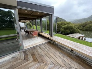 Un espace de restauration extérieur moderne, doté d’une terrasse en bois, de murs en verre et offrant une vue sur un lac et des montagnes.