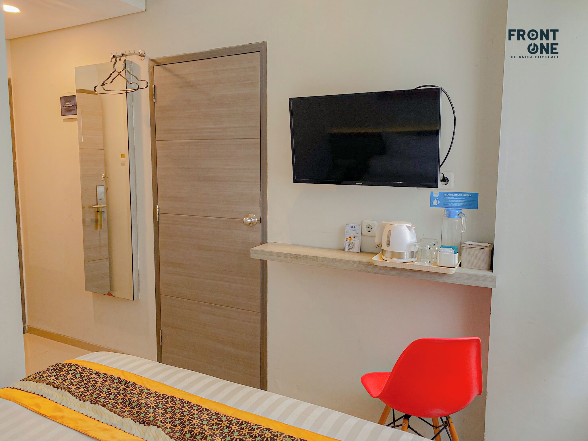 Una camera d'albergo con una porta in legno, una TV montata a parete, un piccolo ripiano con un bollitore, tazze e una sedia rossa.