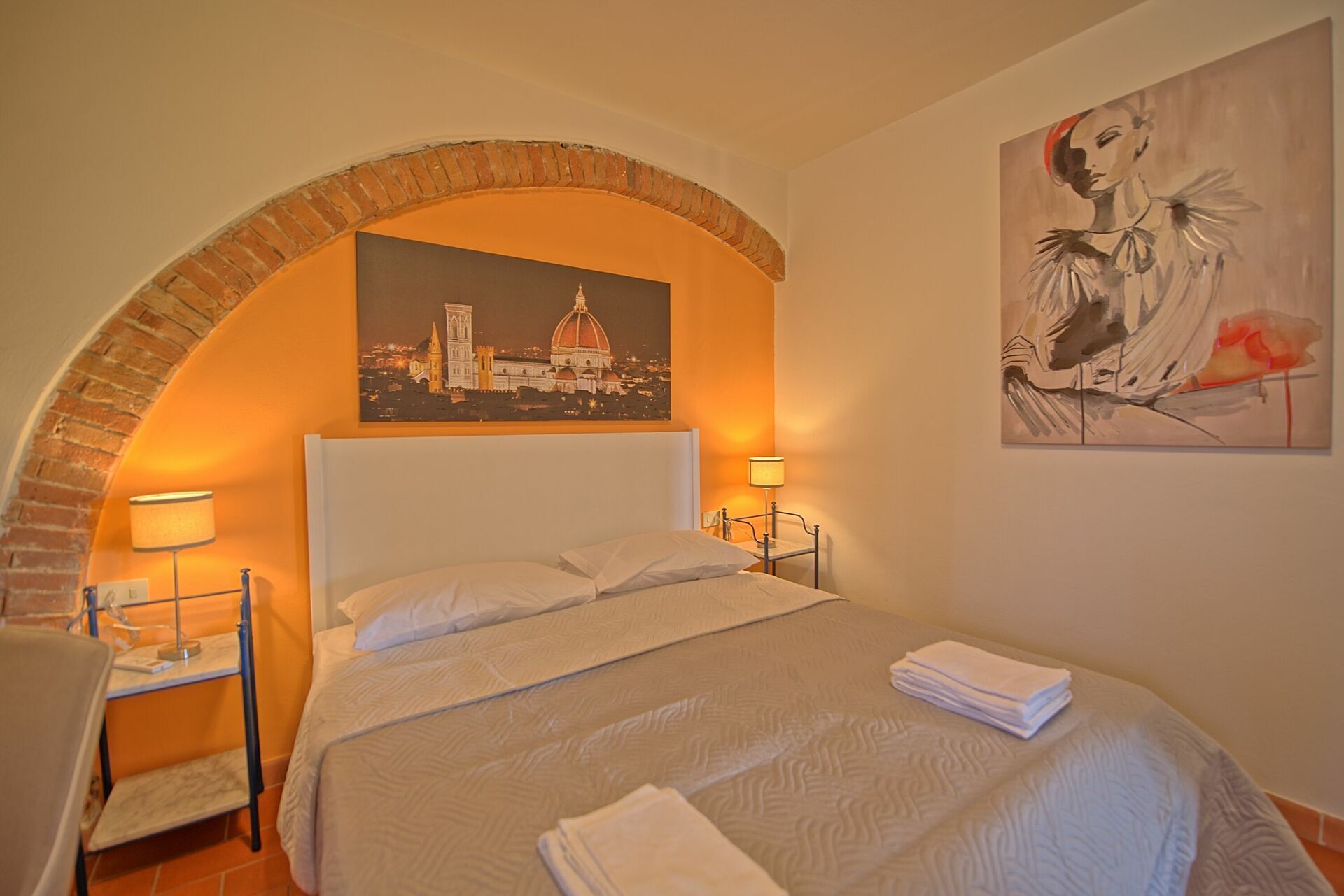 Habitación de hotel con una cama, dos mesitas de noche, una lámpara, un cuadro y una pared de ladrillo en arco.