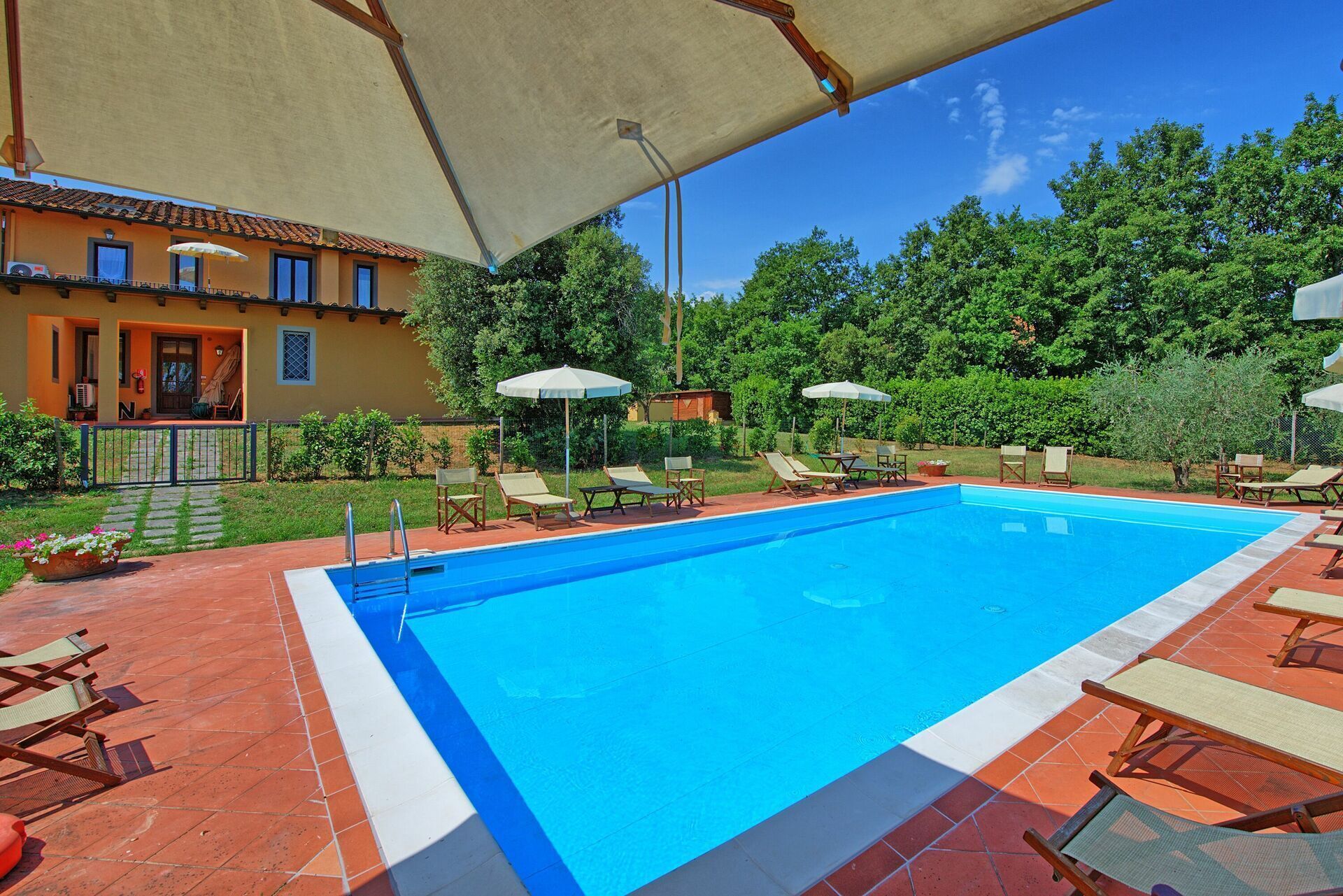 A swimming pool with lounge chairs and umbrellas, surrounded by a garden and a building with a tiled roof.
