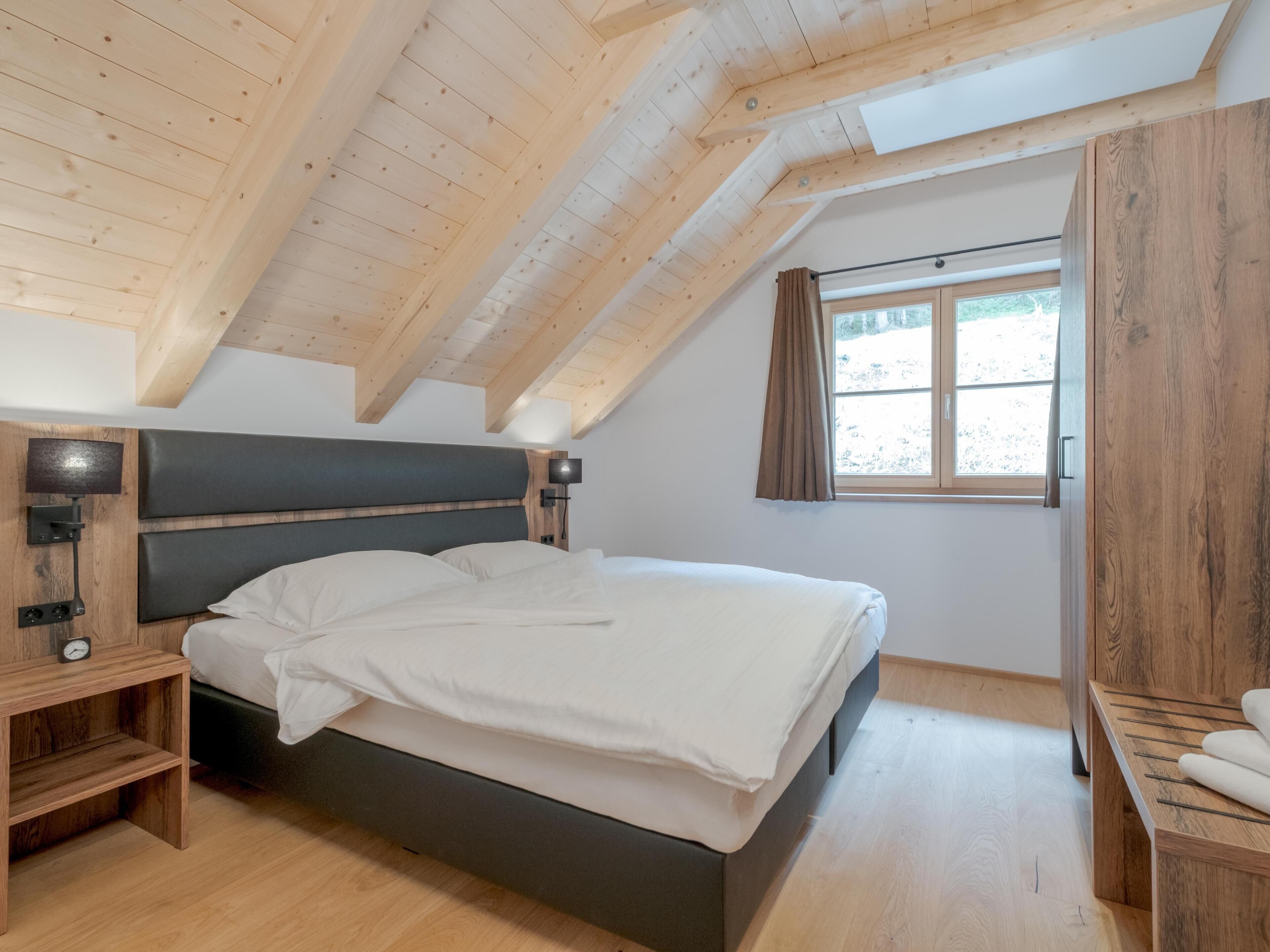 Une chambre avec un plafond en bois, un lit avec une literie blanche, une table de chevet avec une lampe et une fenêtre avec des rideaux.