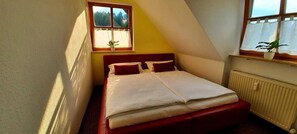 Ein Einzelbett mit Kopfstütze, ein Fenster mit Vorhängen, ein Heizkörper und eine Pflanze auf der Fensterbank.