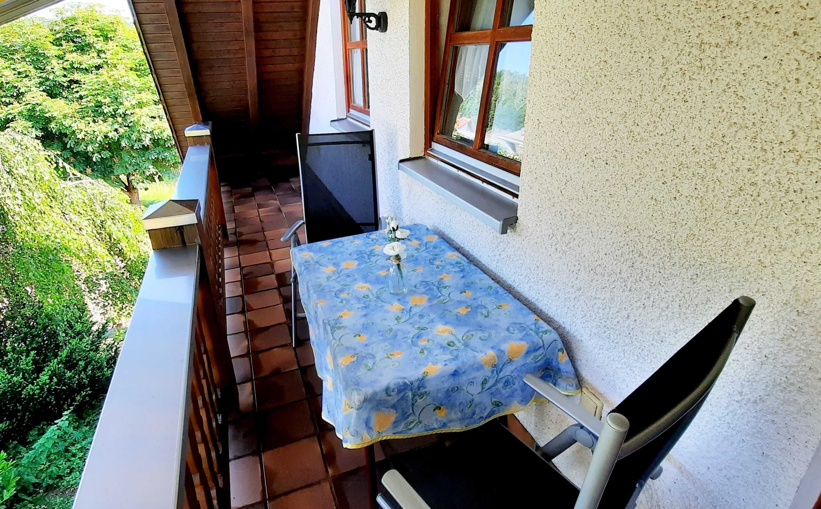 A balcony with a table and chairs, a small table, and a window with a view of greenery.