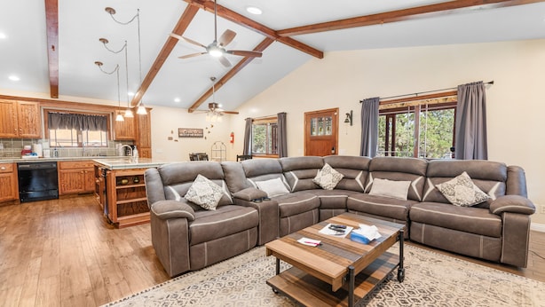 A spacious living room with a large L-shaped sectional sofa, a wooden coffee table, and a kitchen area with wooden cabinets and a window. - Moose Tracks Lodge - Game Room, Ping Pong, Shuffleboard, Barbecue (Big Bear City)