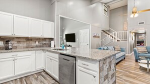 A modern kitchen with white cabinets, a granite countertop, and a dishwasher. There is a staircase leading to a living area with a blue sofa and a ceiling fan.